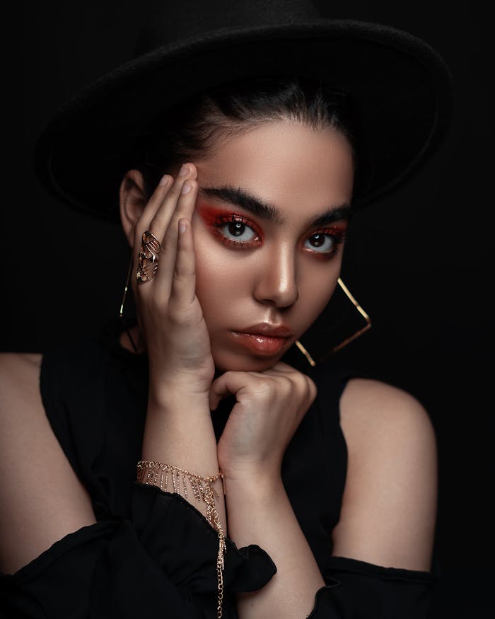 Elegant portrait of a woman with bold makeup, wearing a black hat and jewelry in a studio setting.